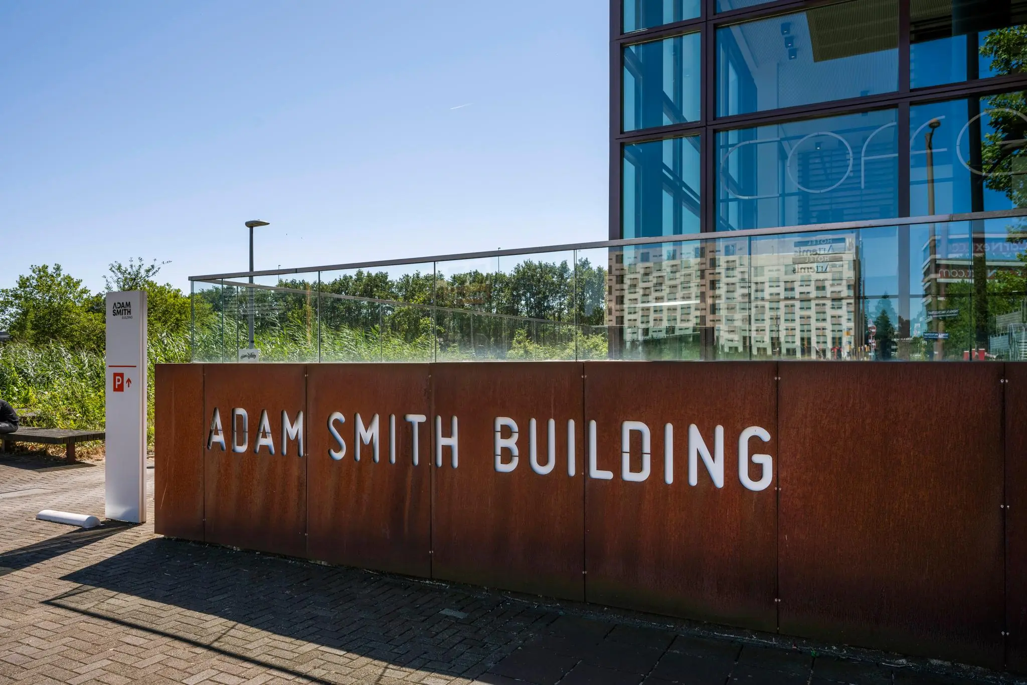 Ingang van het Adam Smith Building met roestkleurige gevel en omliggend groen aan de Thomas R. Malthusstraat.